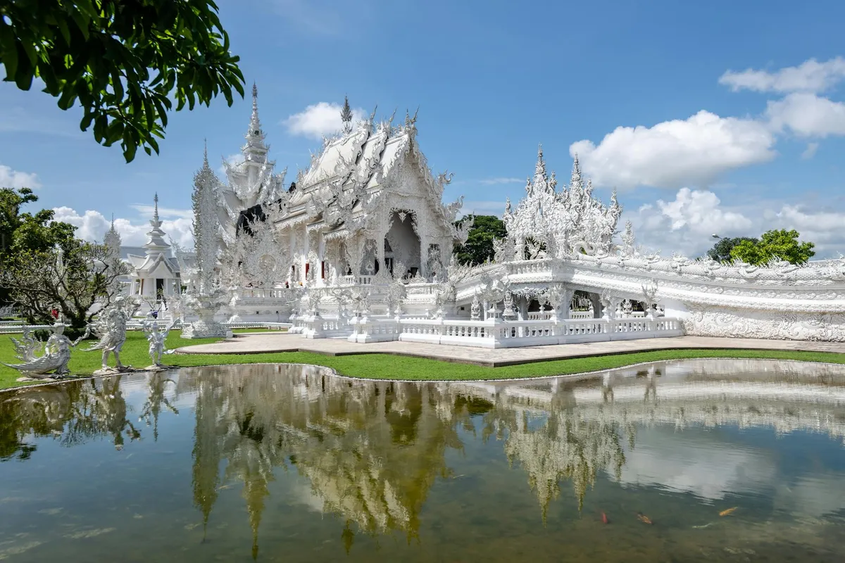 Stunning white architecture of Wat Rong Khun reflected in a tranquil pond under a blue sky in Chiang Rai, Thailand.
